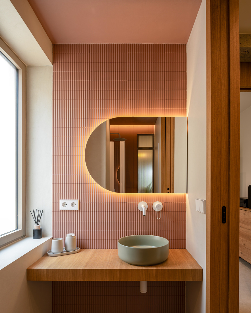 Fotografía de baño con revestimiento cerámico rosa Fotografía de arquitectura interior del baño de Casa Melón, con revestimiento cerámico rosa, lavabo verde y espejo retroiluminado sobre encimera de madera.