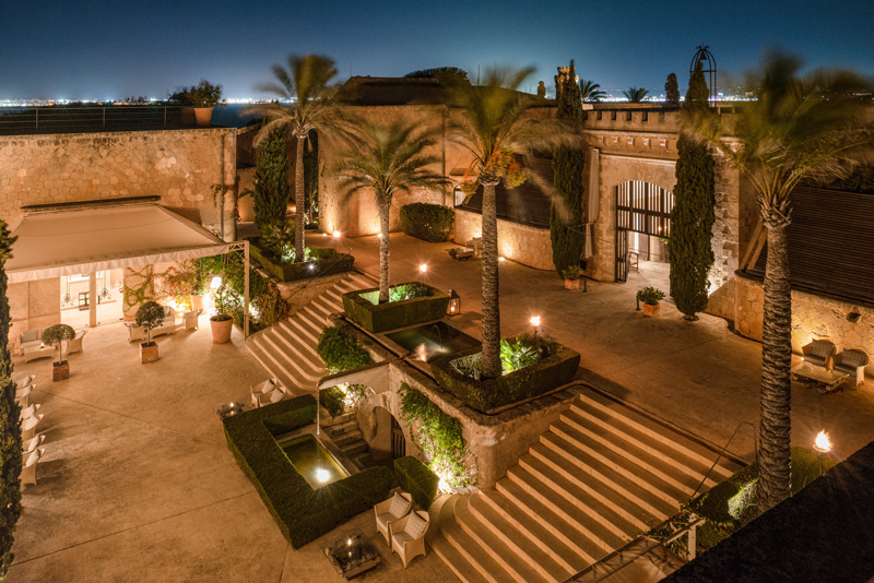Vista elevada del patio de Cap Rocat iluminado de noche, con escaleras y palmeras